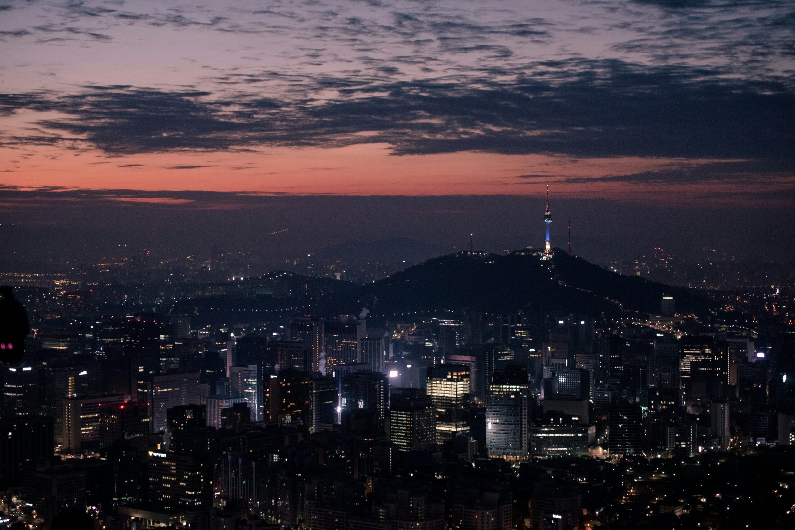 Seoul city skyline at night with Namsan Seoul Tower glowing on a mountain under a dark evening sky.