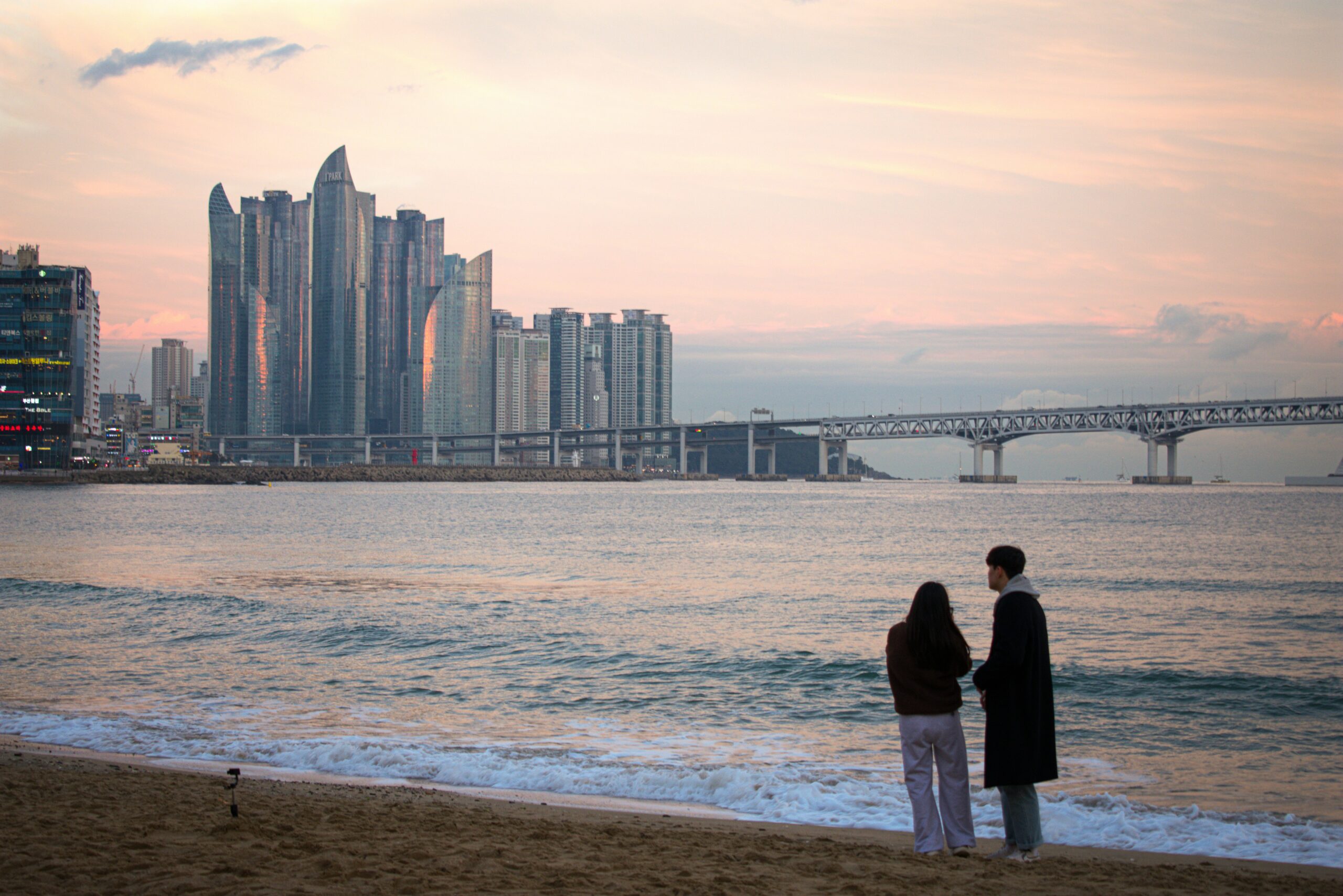 Two people standing on a beach in Busan at sunset, overlooking the sea, city skyline, and Gwangan Bridge.