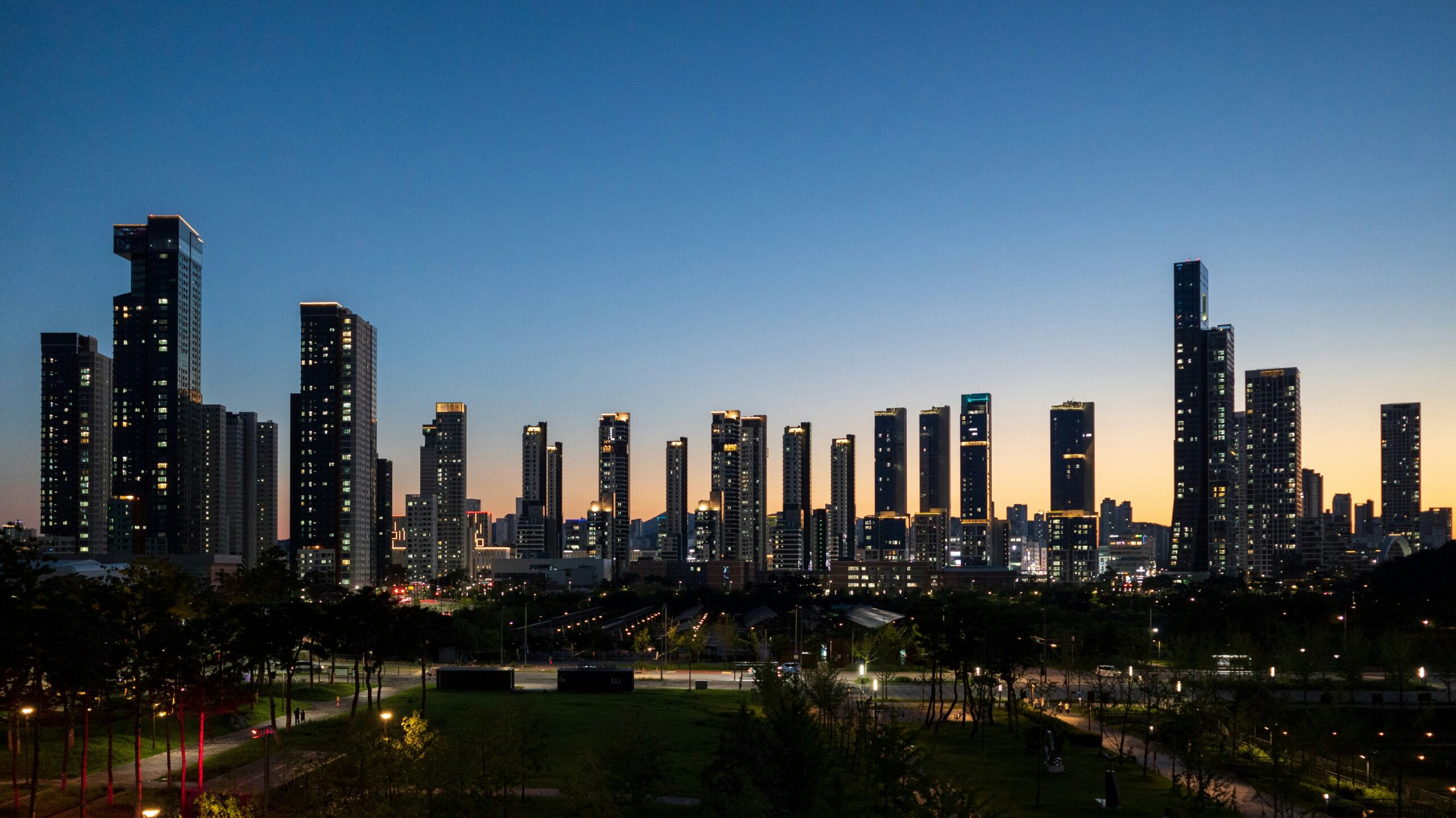 Modern South Korean city skyline at dusk with high-rise buildings, parkland, and illuminated streets.