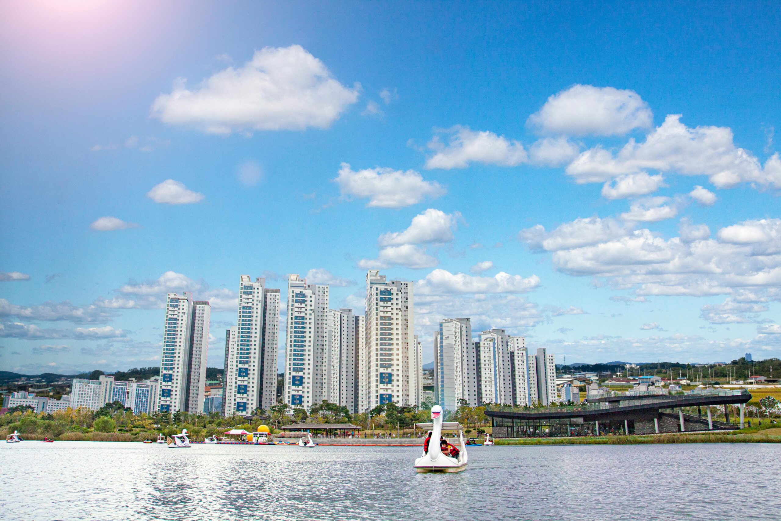 Daegu city lake with swan boats, apartment towers, blue sky, and white clouds.
