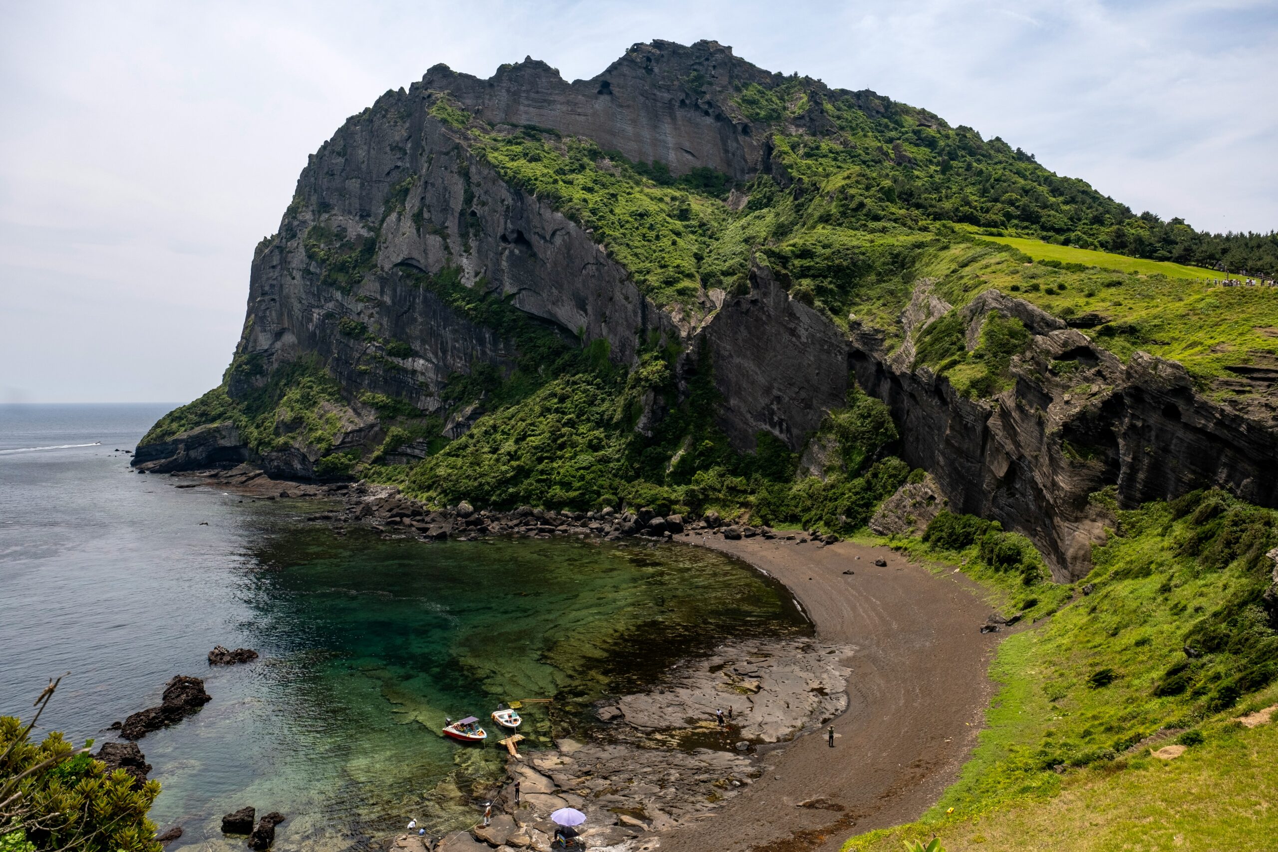 Jeju Island coastal cliff with green volcanic rock formations, clear seawater, and a quiet beach.
